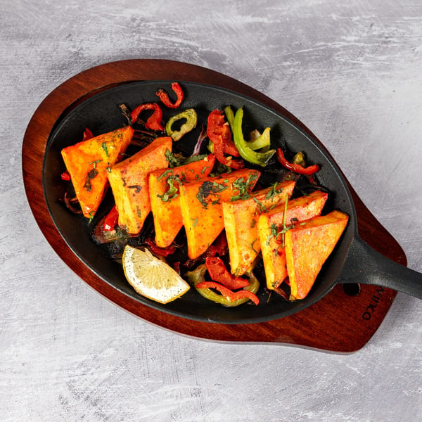 Triangular paneer pieces with bell peppers and a lemon wedge in a sizzling cast iron pan.