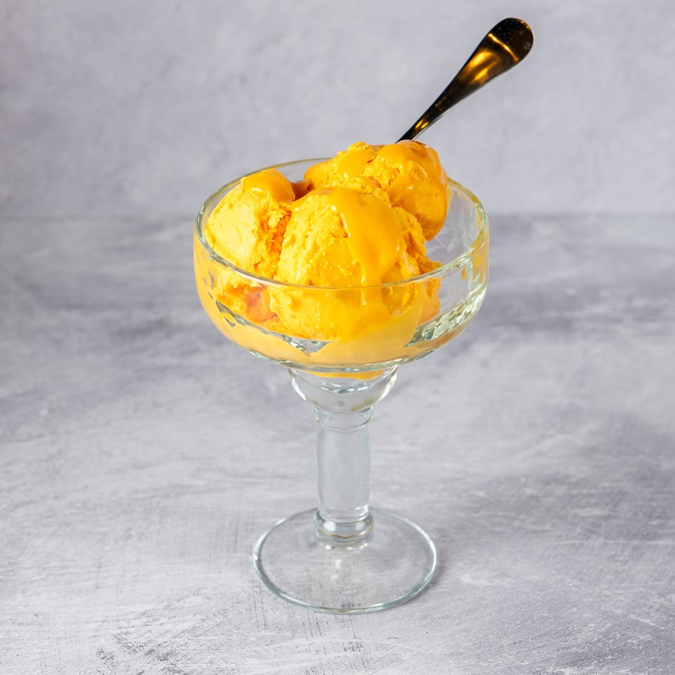 Three scoops of bright orange mango ice cream in a clear glass dessert cup with a spoon.
